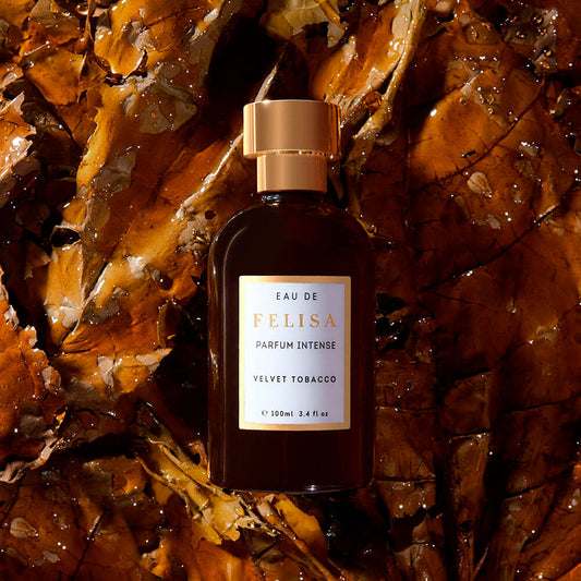 Frasco de Velvet Tobacco da Felisa sobre folhas de tabaco secas e úmidas, com luz quente e atmosfera noturna