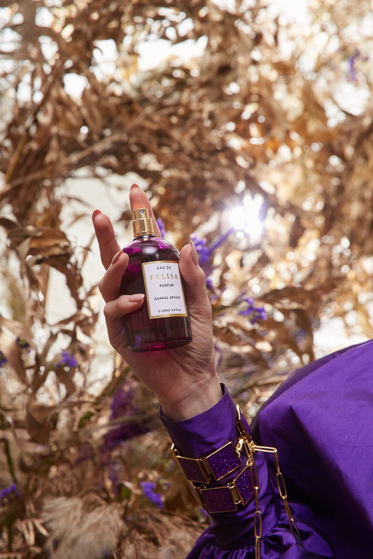 mão segurando o frasco roxo de Sandal Affair ao ar livre, entre galhos secos e flores roxas, com luz de fundo do entardecer - perfume Sandal Affair, Felisa Beauty, perfumaria de nicho brasileira de luxo